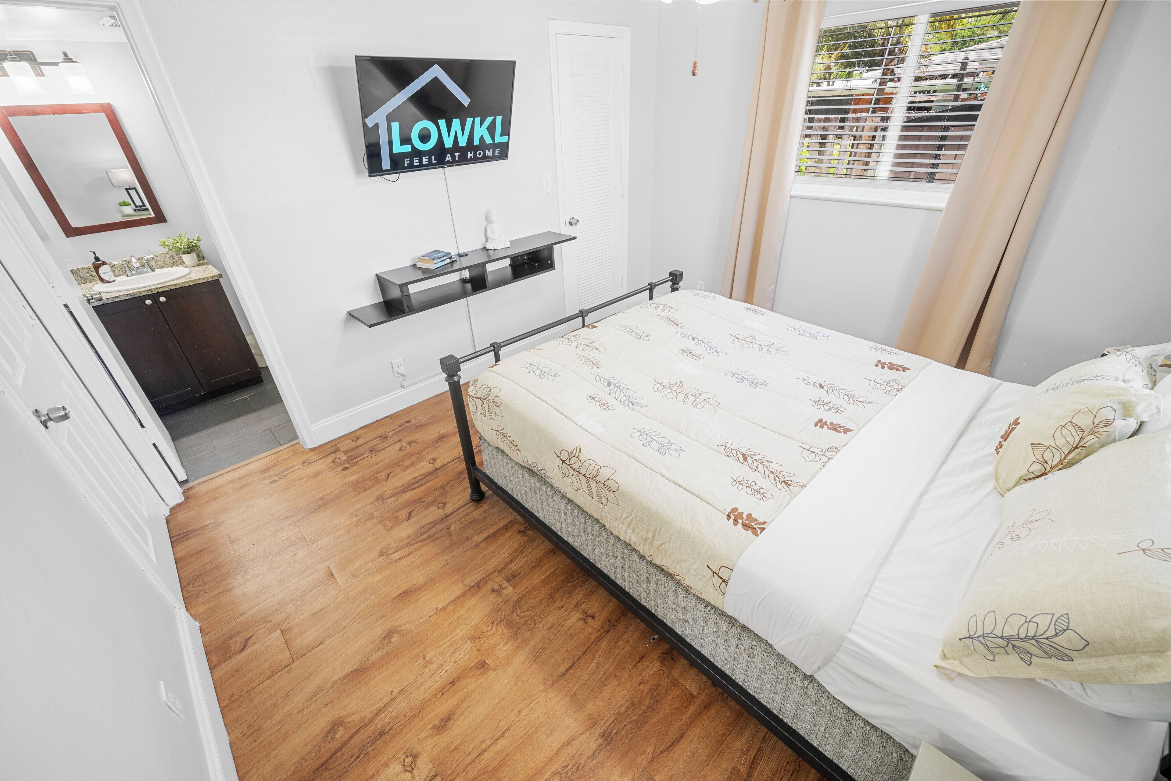 Une chambre à coucher avec un lit, un téléviseur fixé au mur, une fenêtre avec des rideaux et une petite étagère sur laquelle se trouve une plante.