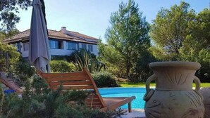 Zona de piscina con una terraza de madera, una planta grande en maceta y una casa al fondo.