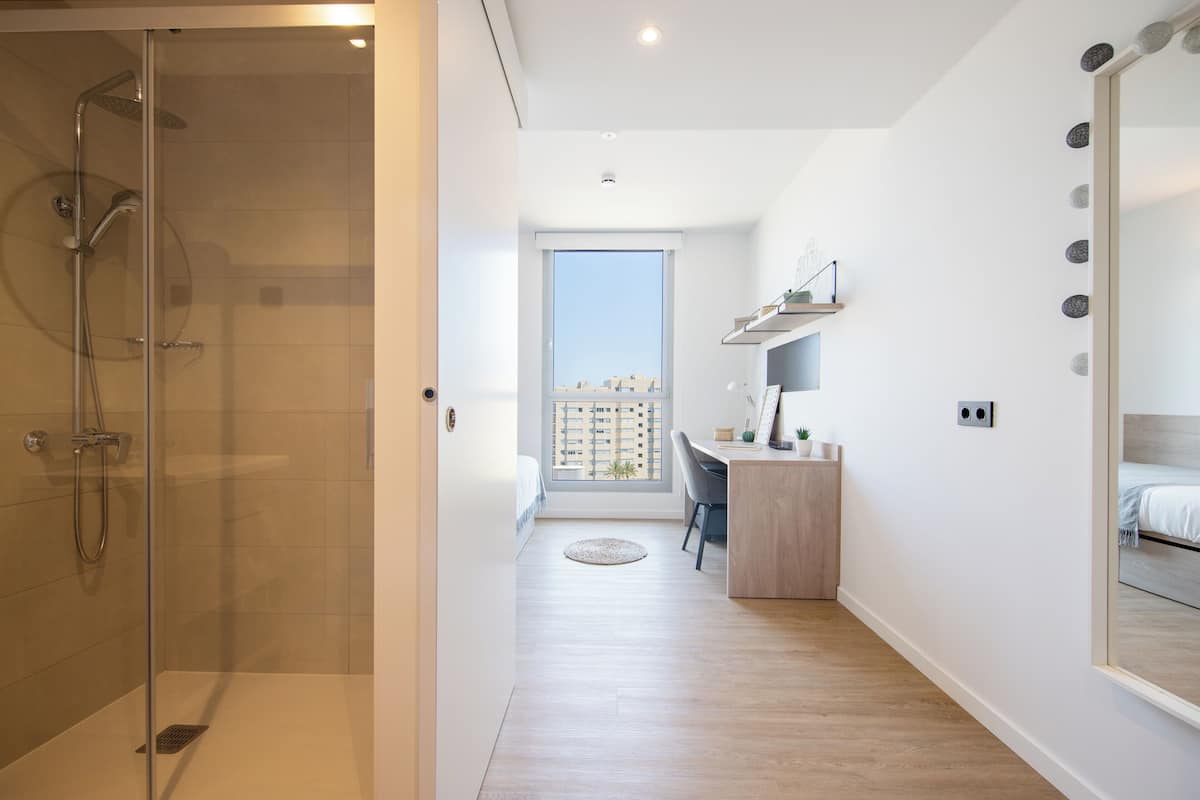 A modern hotel room with a shower, a desk, a chair, and a large mirror.