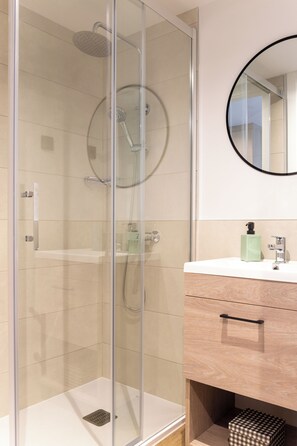A modern bathroom with a glass-enclosed shower, a wooden vanity with a mirror, and a small checkered box on the floor. - Micampus Barcelona Student Residence (Sant Adria de Besos)