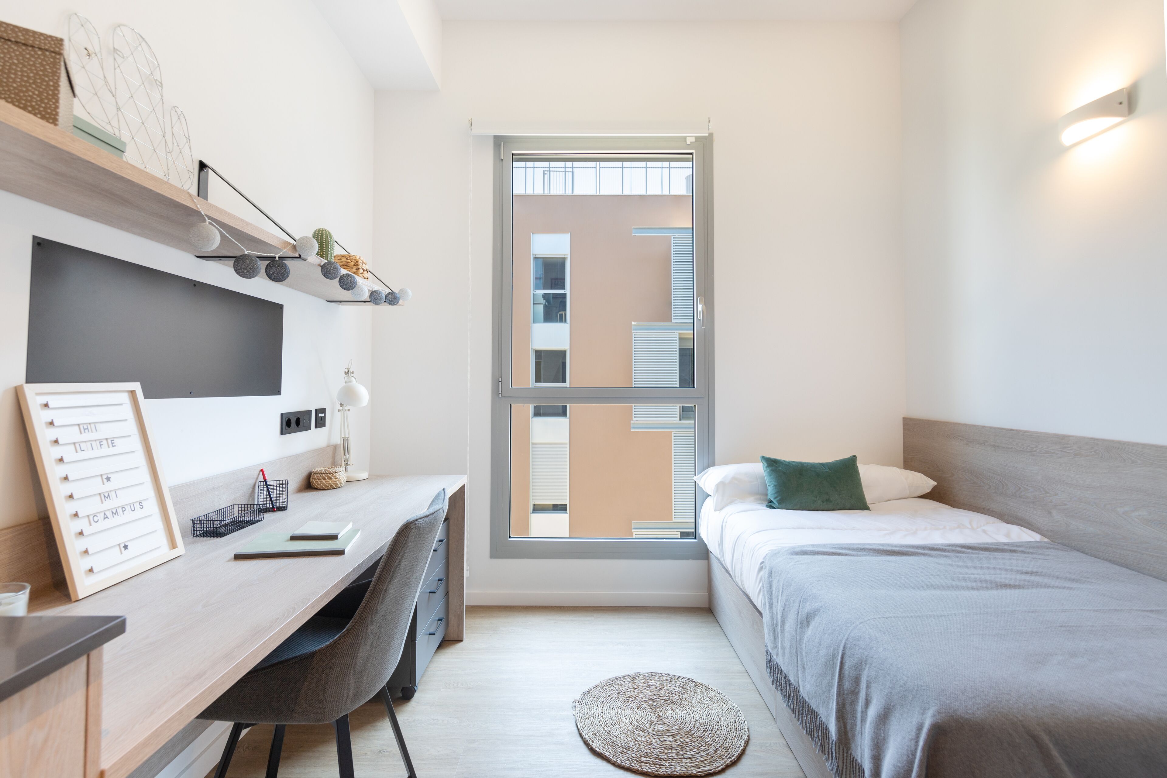 Ein kleines Schlafzimmer mit Schreibtisch, Bett und Fenster, mit Blick auf ein Gebäude.