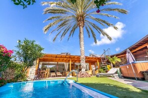A poolside area with a palm tree, a wooden deck, and a building with large windows. - Anfi Tauro, Private Pool, Hot Tub in pretty garden (Mogán, Gran Canaria)