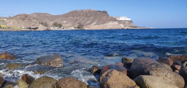 A rocky shoreline with large boulders and a distant view of a fortified hill with buildings. - Anfi Tauro, Private Pool, Hot Tub in pretty garden (Mogán, Gran Canaria)