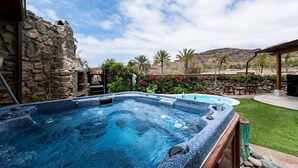 Een hottub met een achtergrond van een stenen muur, omgeven door een terras met tuinmeubels en weelderige beplanting.