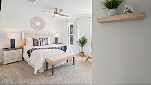 A bedroom with a bed, bedside tables, a bench, a ceiling fan, a window with curtains, and a wall-mounted shelf with plants.