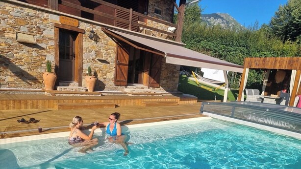 A swimming pool with two people in the water, surrounded by stone buildings and wooden deck.