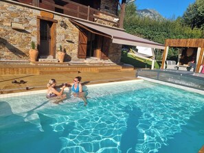 Une piscine où se trouvent deux personnes, entourée de bâtiments en pierre et d’une terrasse en bois.