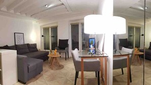 Un salón moderno con un sofá gris, una mesa de comedor de cristal y un espejo grande que refleja la estancia.