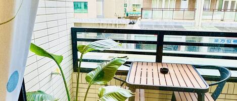 A balcony with a wooden table and chairs, potted plants, and a view of a cityscape with buildings.