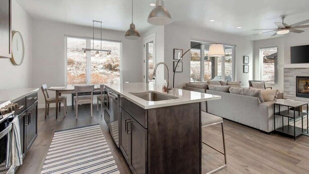 A modern kitchen with a central island, a dining area with a table and chairs, a living room with a sofa and a TV, and large windows with a view of the outdoors.