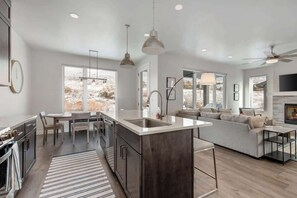 A modern kitchen with a central island, a dining area with a table and chairs, a living room with a sofa and a TV, and large windows with a view of the outdoors. - Hideout Hideaway - 4BR Townhome w/ Private Hot Tub (Daniel)