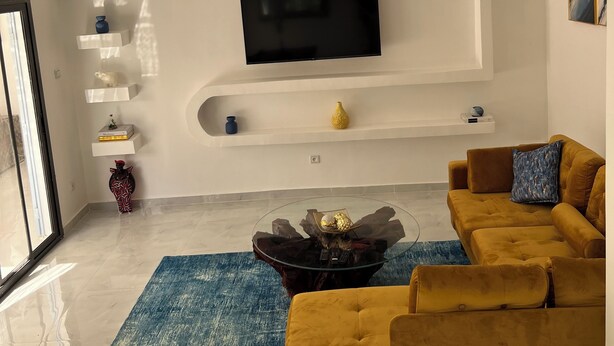Un salon moderne avec un téléviseur à écran plat fixé à un mur blanc, un canapé de couleur moutarde, un tapis bleu et une table basse en verre agrémentée d’objets décoratifs.