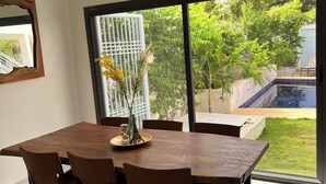 Un coin repas avec une grande table et des chaises, un petit pouf rond et une vue sur un jardin et une piscine, accessible par une porte-fenêtre coulissante.