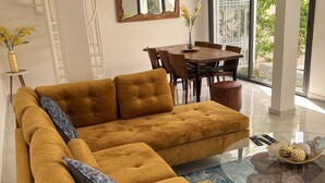 Un salon moderne avec un canapé d’angle de couleur moutarde, une table à manger avec des chaises, une table basse en verre et un grand miroir au mur.