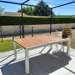 Ein Holztisch mit weißen Beinen, ein Korbstuhl und ein Metallzaun in einem Garten.