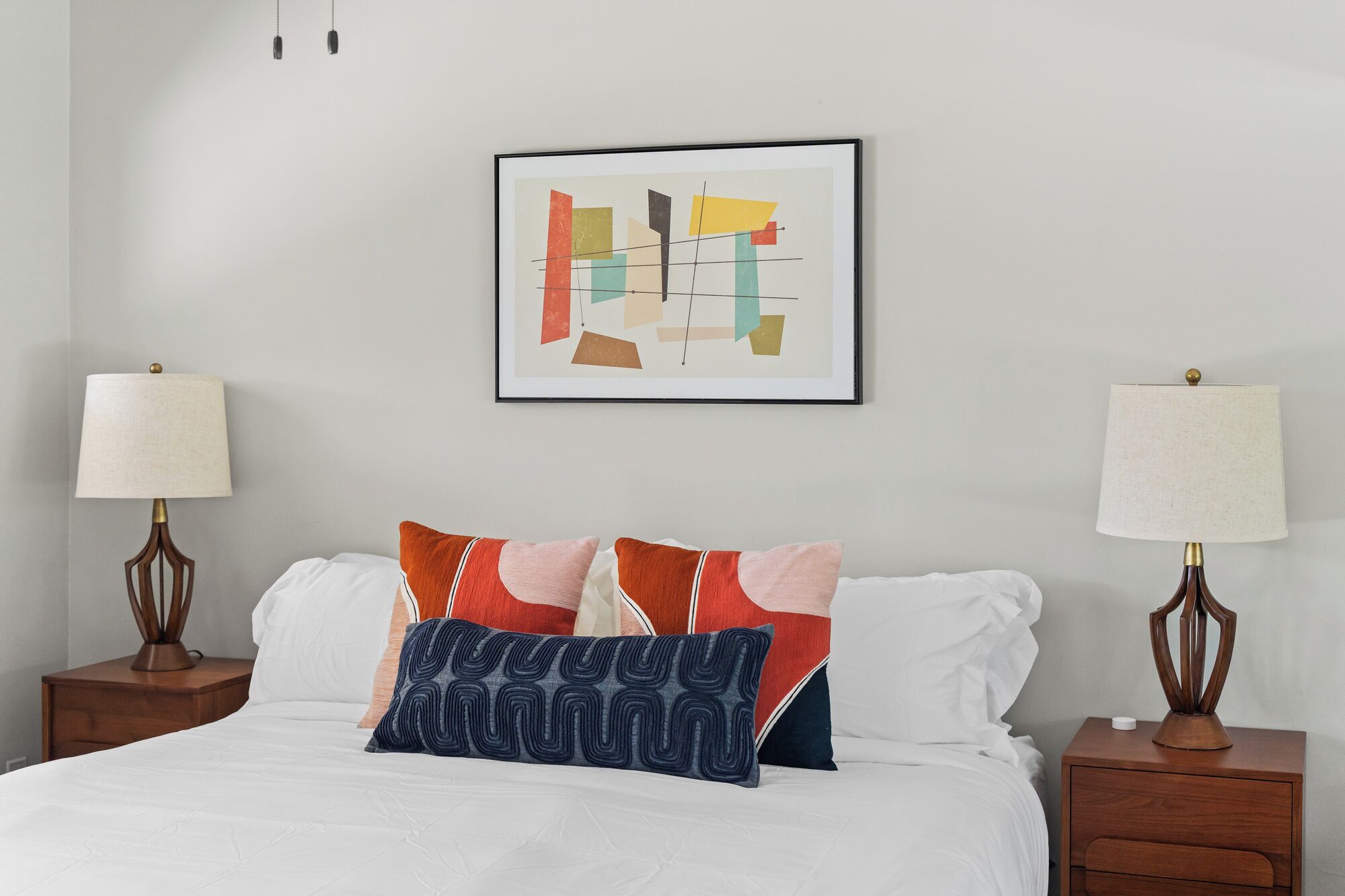 A neatly made bed with white bedding and multiple pillows, two bedside tables with lamps, and a framed abstract artwork on the wall.