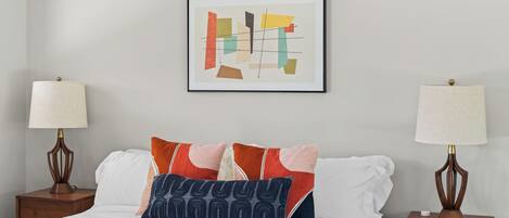 A neatly made bed with white bedding and multiple pillows, two bedside tables with lamps, and a framed abstract artwork on the wall.