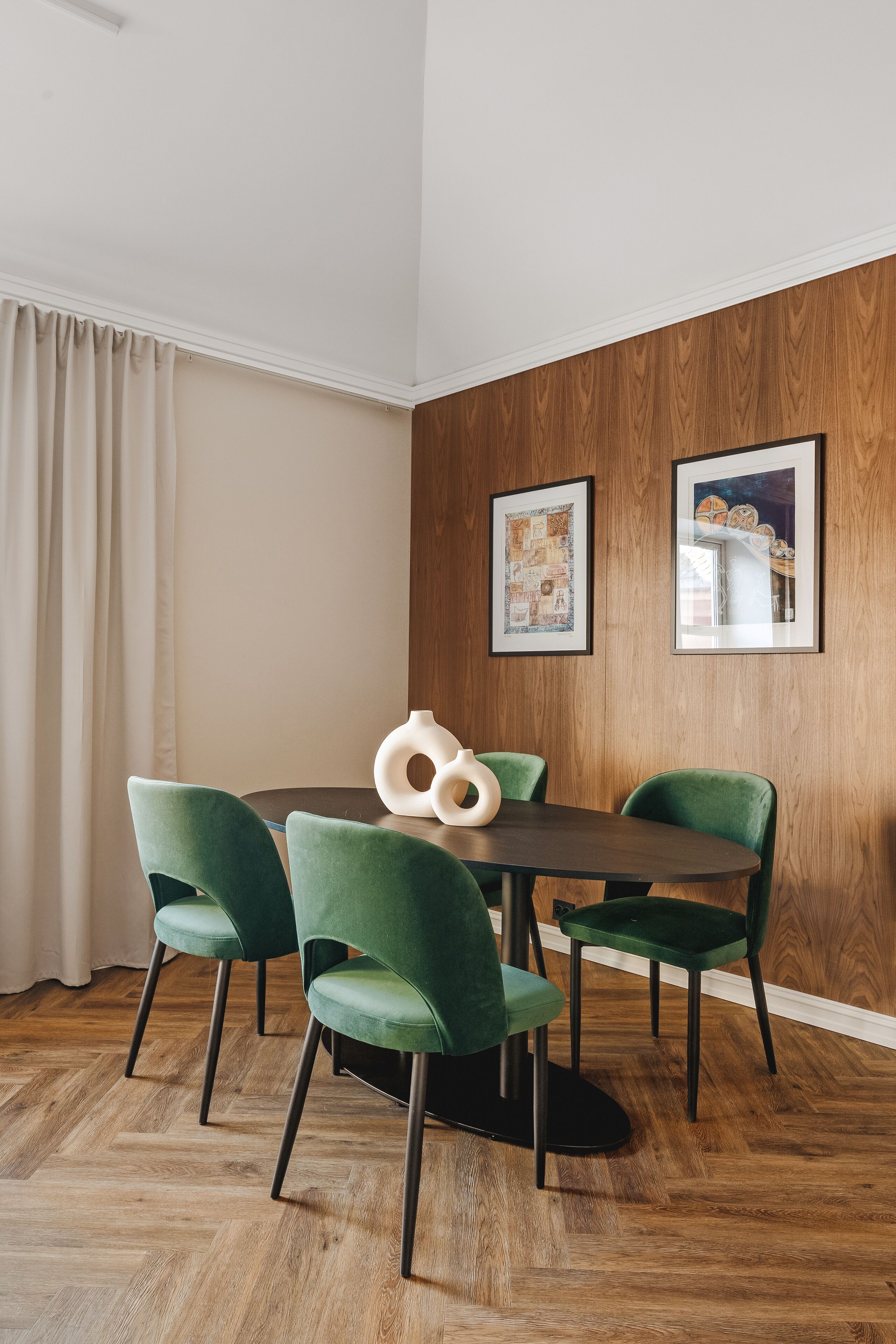 Un comedor con una mesa redonda, cuatro sillas verdes, paneles de madera y dos obras de arte enmarcadas.