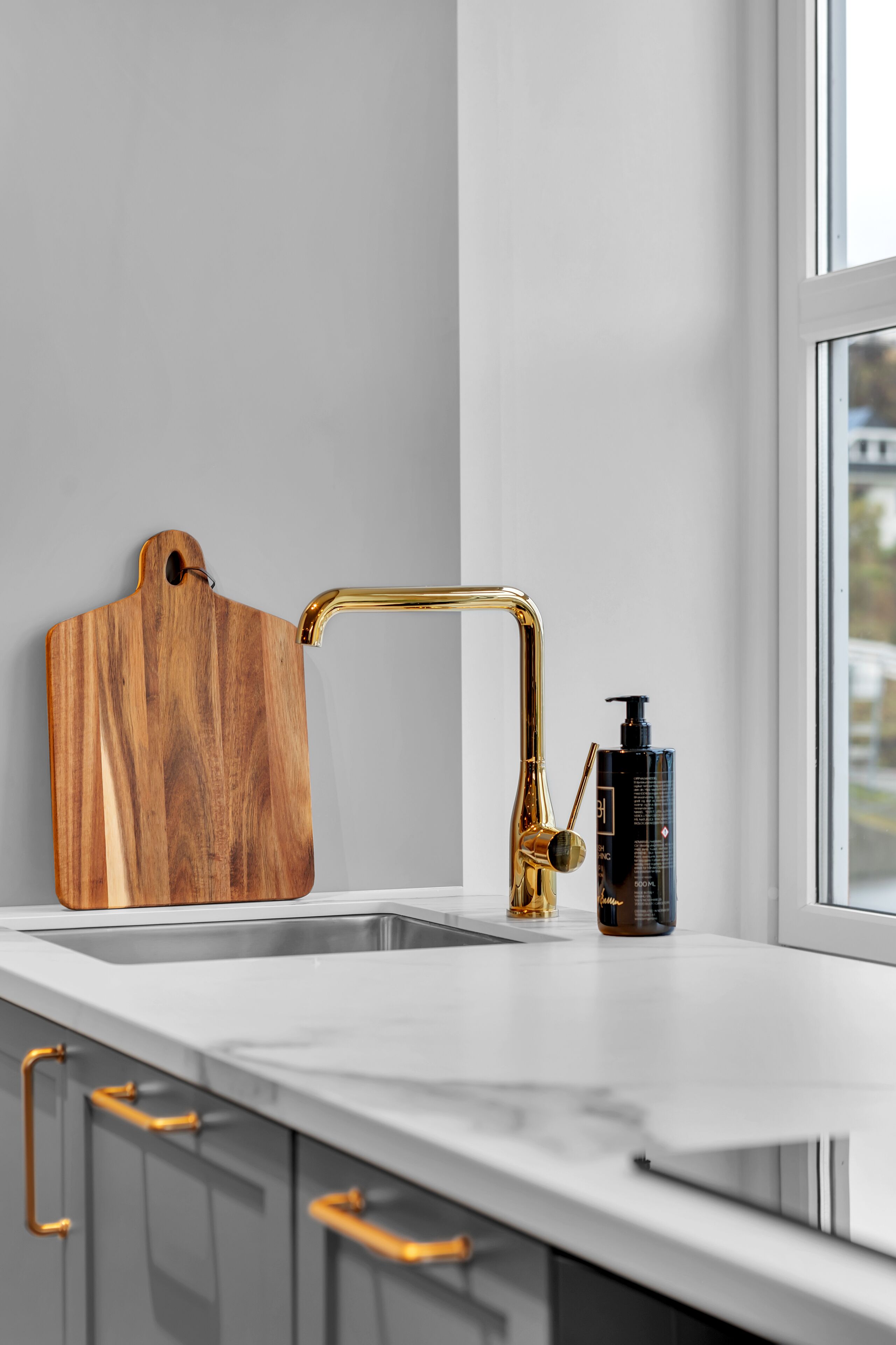 Een keuken met een wit aanrecht, een houten snijplank, een gouden kraan en een zeepfles.