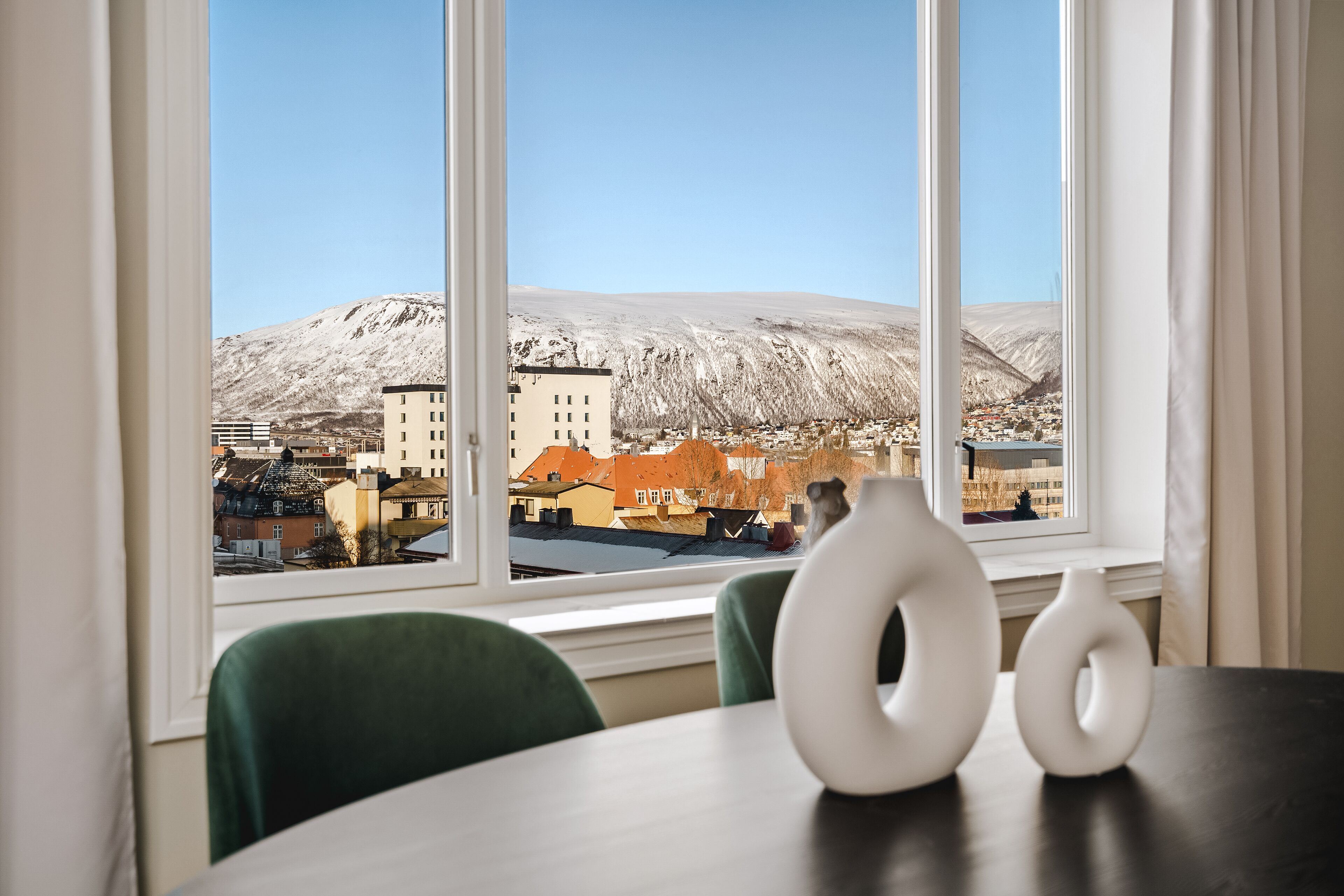 A room with a view of a snowy mountain and buildings. There are two white vases on a table.