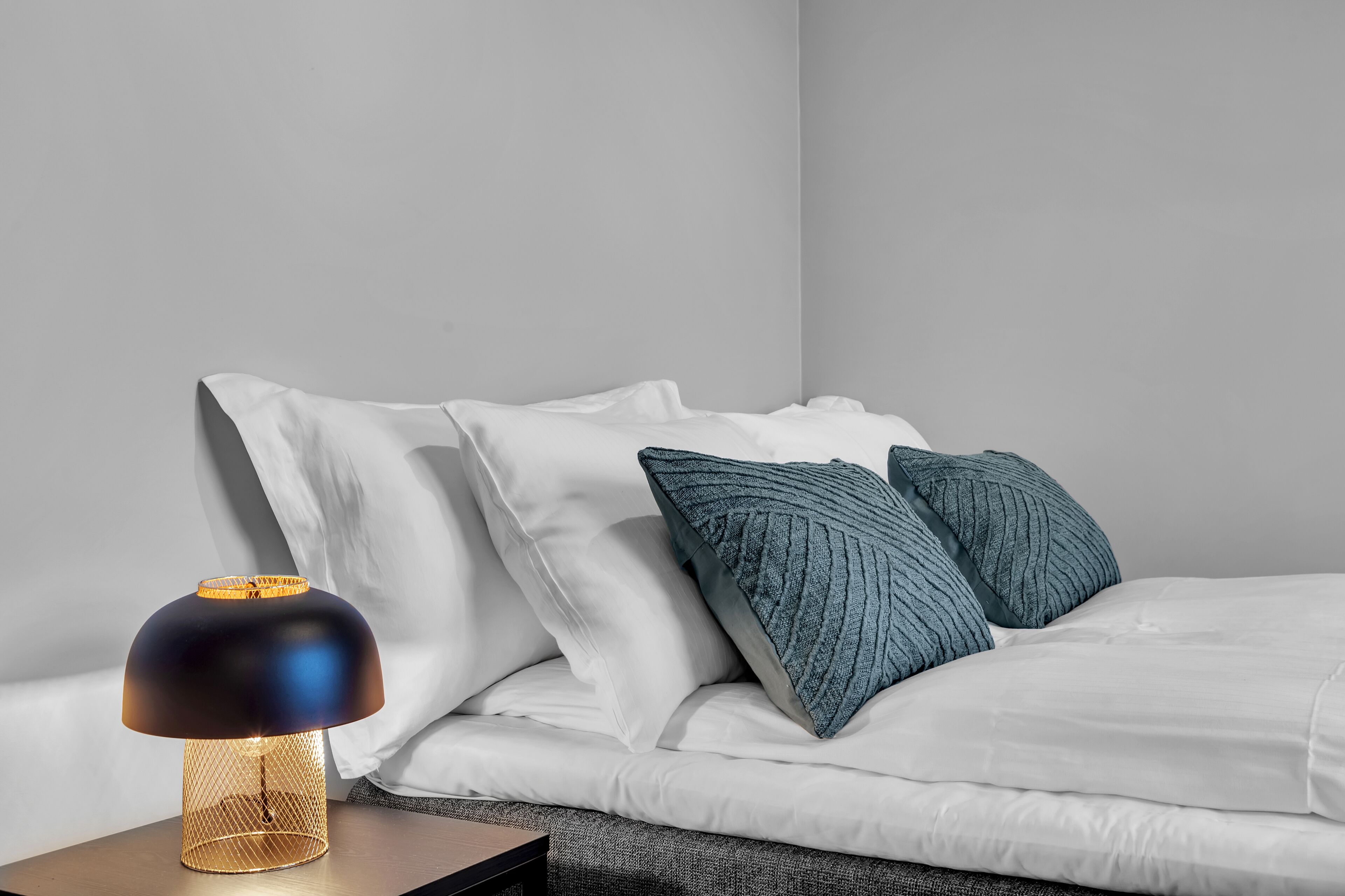 Un lit bien fait, avec une literie blanche et deux oreillers bleus, une lampe de table moderne avec un pied doré, et un mur sobre en arrière-plan.