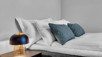 Un lit bien fait, avec une literie blanche et deux oreillers bleus, une lampe de table moderne avec un pied doré, et un mur sobre en arrière-plan.