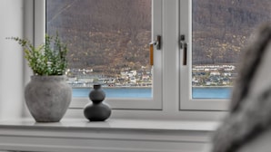 Et soveværelse med et stort vindue der udover et bjergrigt landskab og et vandområde.