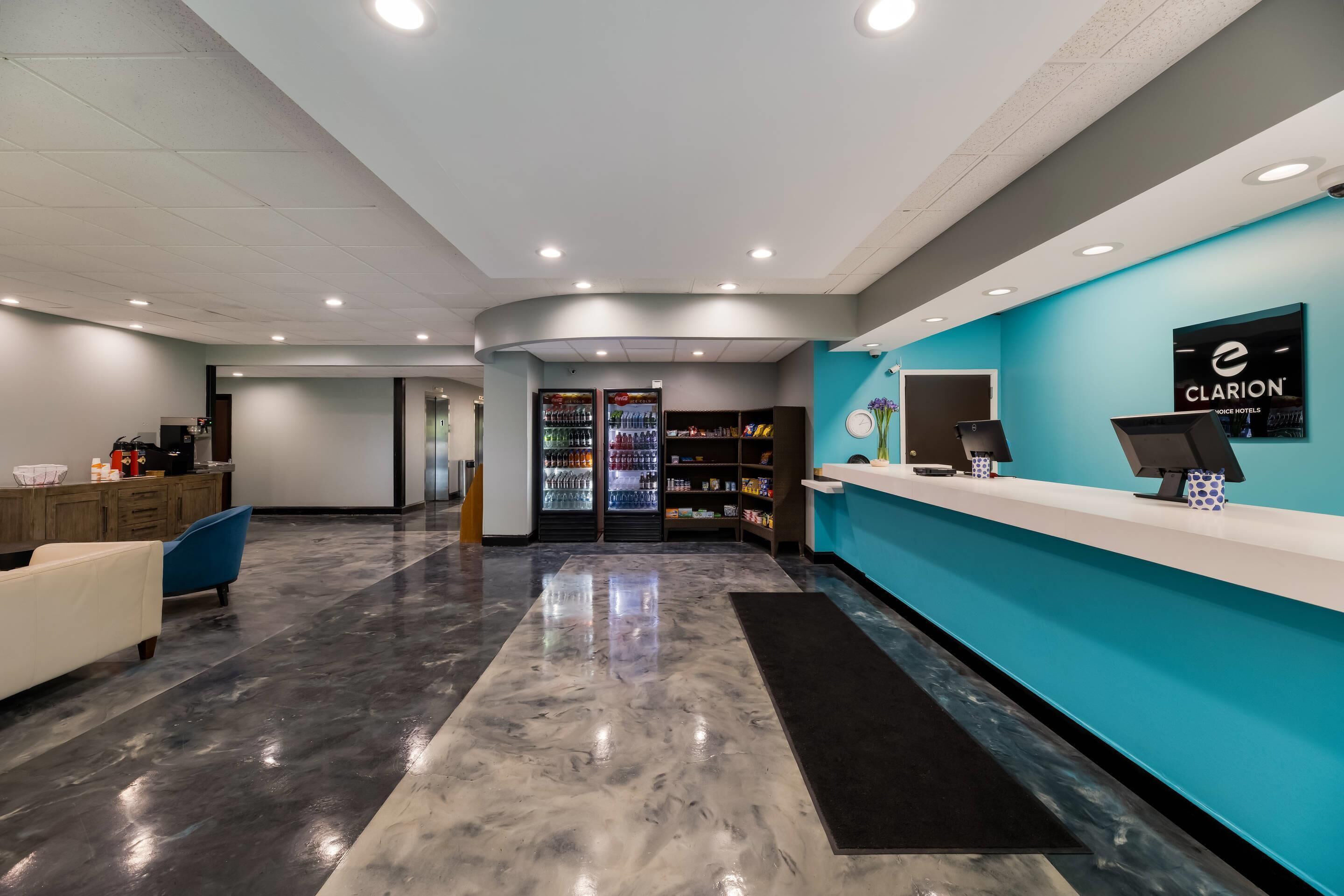 A hotel reception area with a blue wall, a reception desk, a refrigerator stocked with beverages, and a seating area with a blue chair and a white sofa.