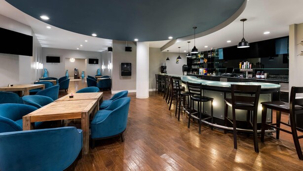 Zona de bar moderna con mesas de madera y sillas azules, techo curvo y una cocina con taburetes de bar.
