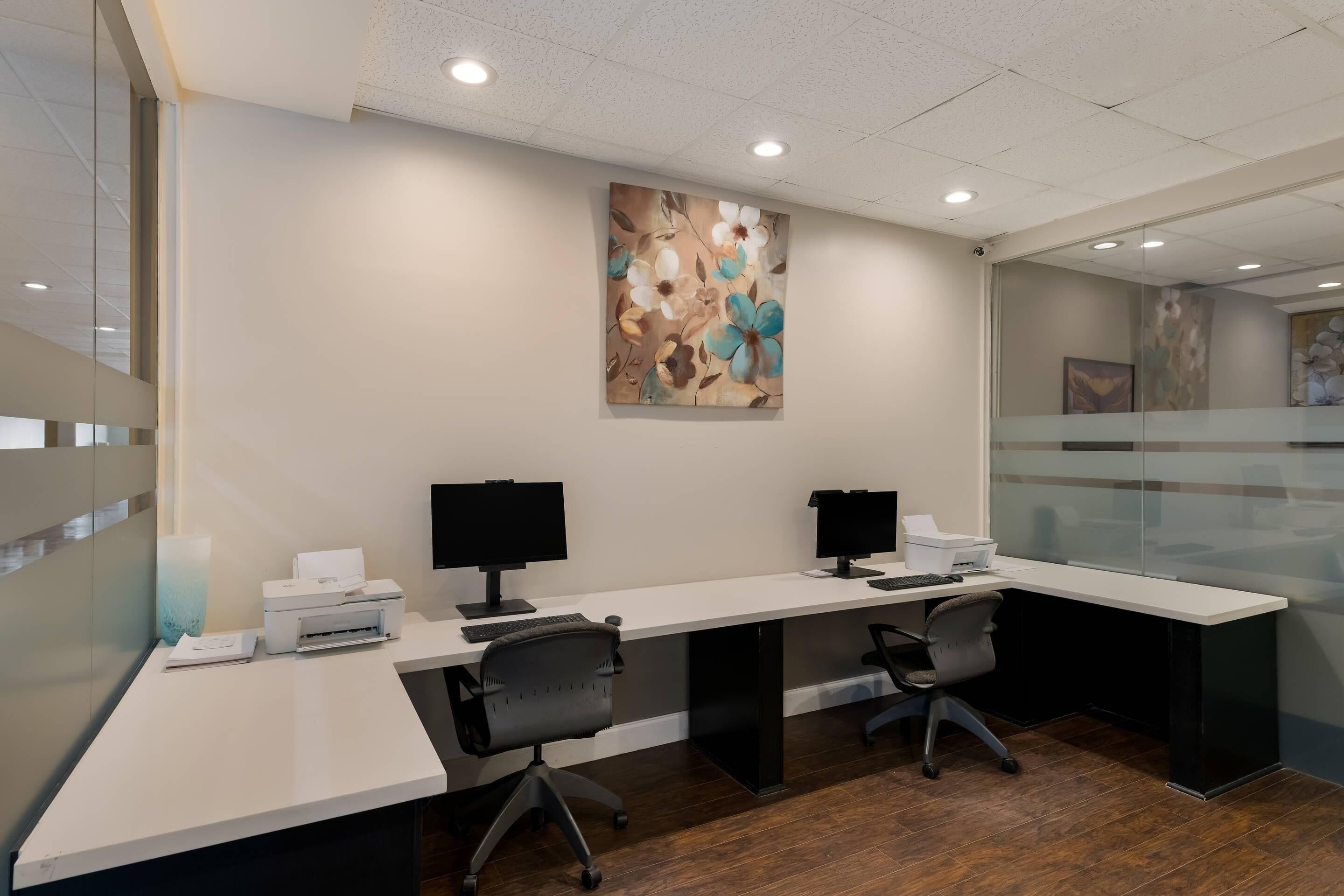 A modern office space with two desks, each equipped with a computer monitor, keyboard, and printer.