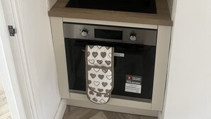 A compact kitchen with a built-in stove and oven, a white tiled backsplash, and a small window with a heart-patterned towel hanging. - Modern welsh cottage (penygraig)