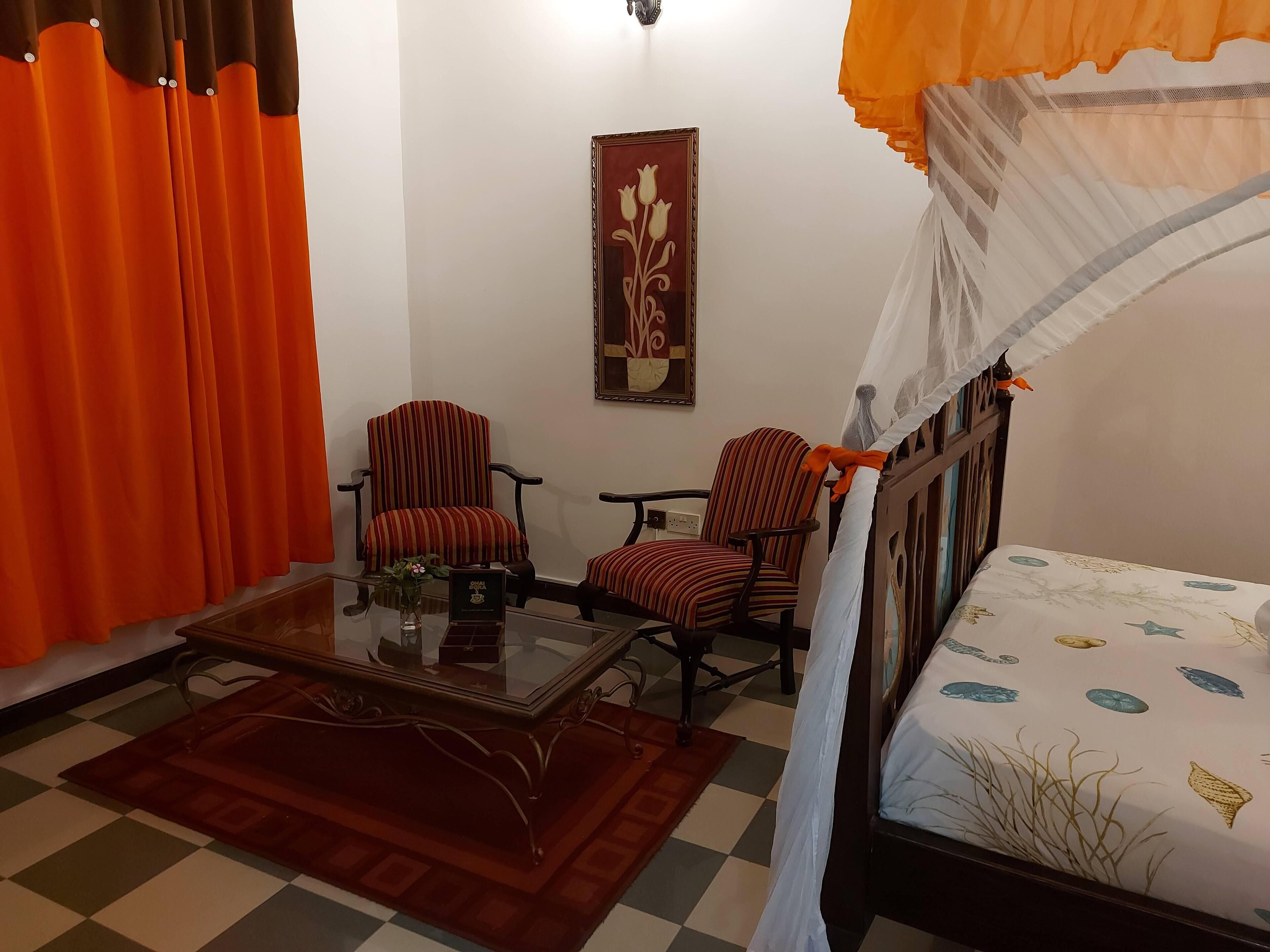 A room with a bed, two striped chairs, a glass coffee table, a framed picture on the wall, and orange curtains.