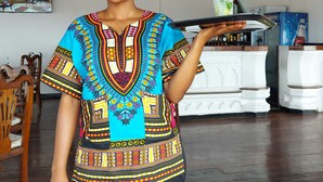 A person in a colorful traditional garment holding a tray in a room with wooden flooring, a bar area, and a painting of a cityscape. - Horizon Palace (Zanzibar Town)