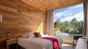 A modern bedroom with a wooden ceiling, a large bed with white bedding and a red throw, a bedside table with a lamp, and a view of greenery through a large window. - Mawün Lodge (Villarrica)