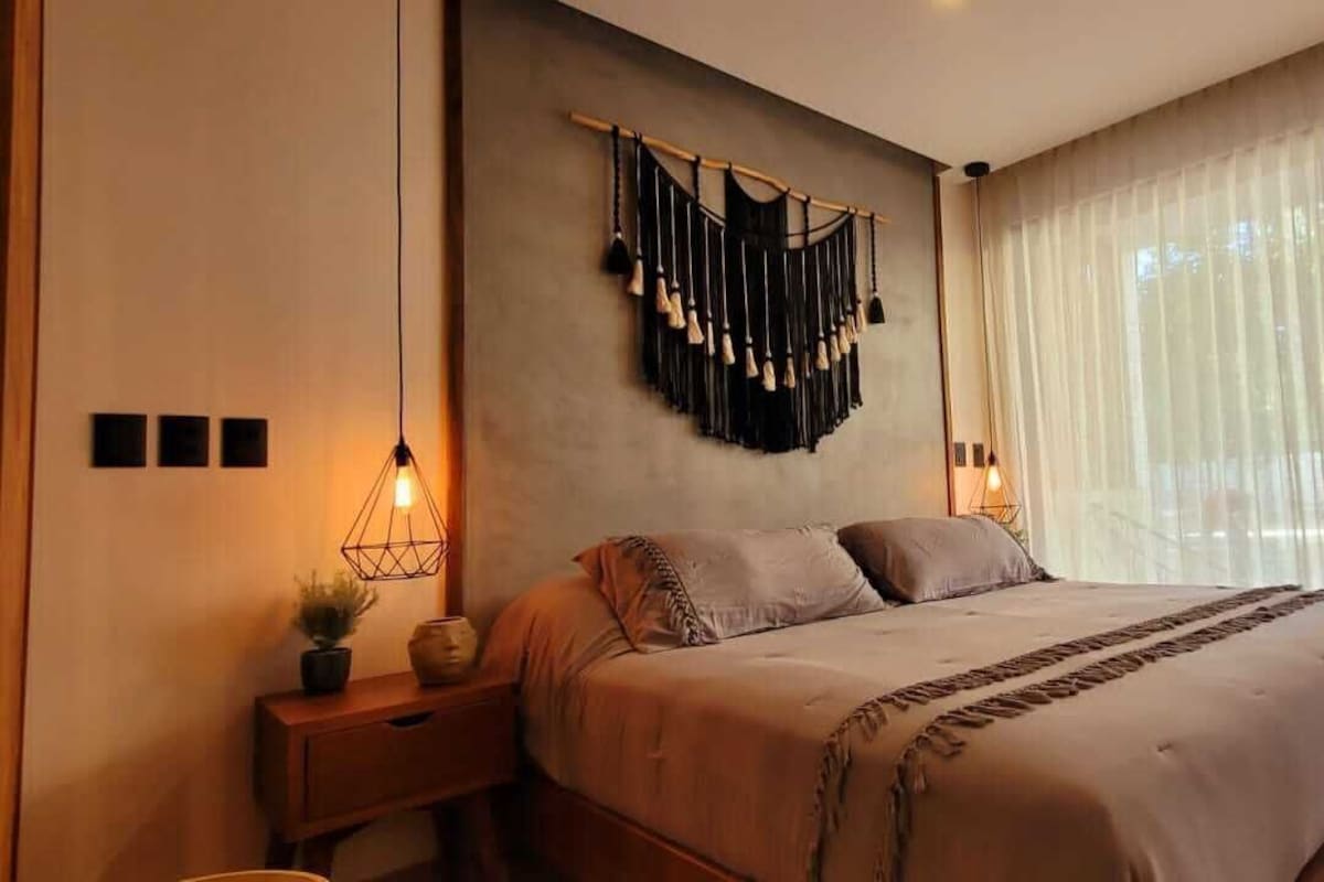 Une chambre à coucher avec un lit, des tables de chevet, une décoration murale en macramé et des rideaux en voile.