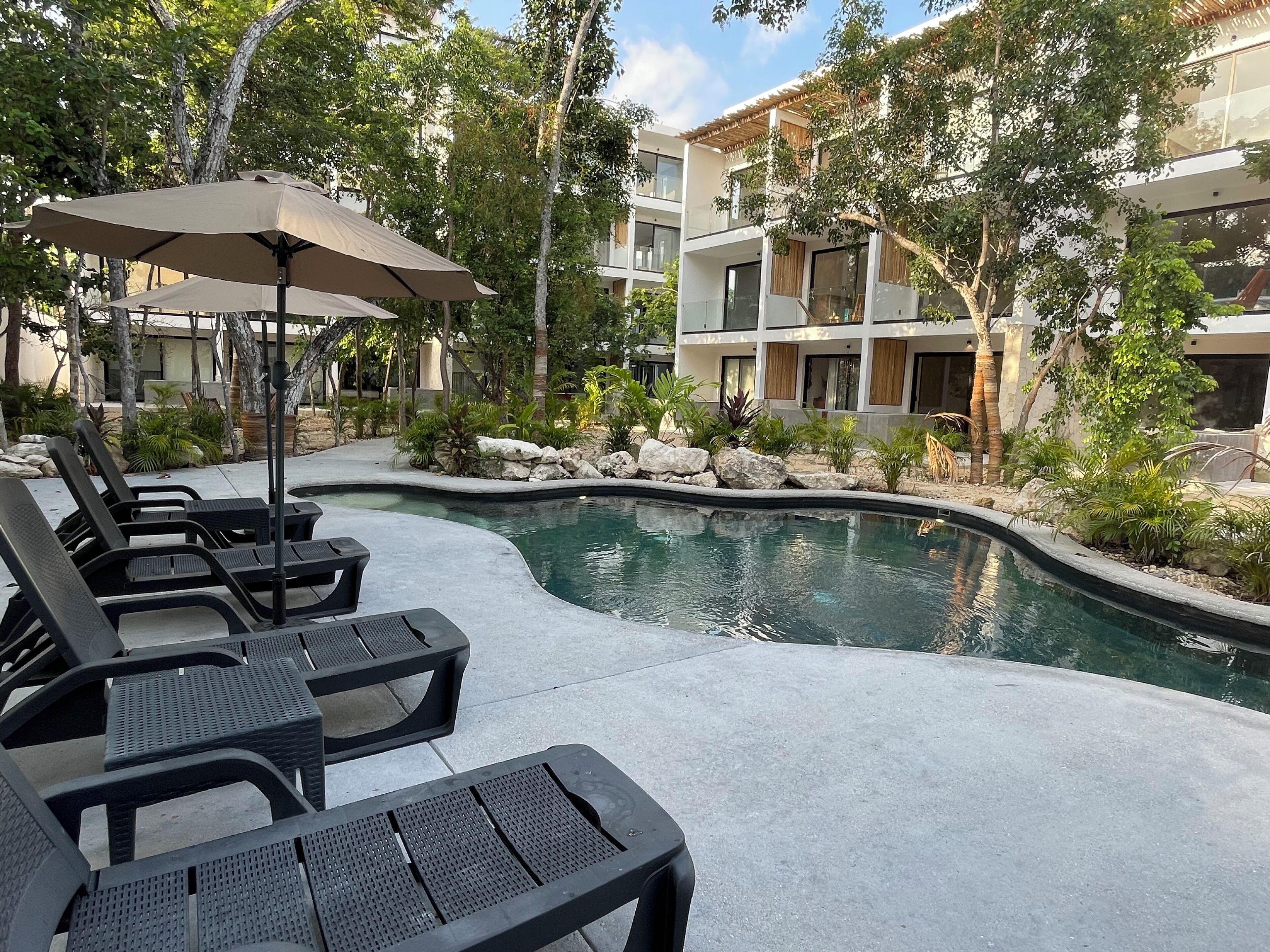 A pool area with lounge chairs and umbrellas, surrounded by greenery and modern buildings.