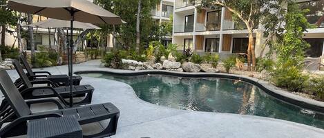 A pool area with lounge chairs and umbrellas, surrounded by greenery and modern buildings.