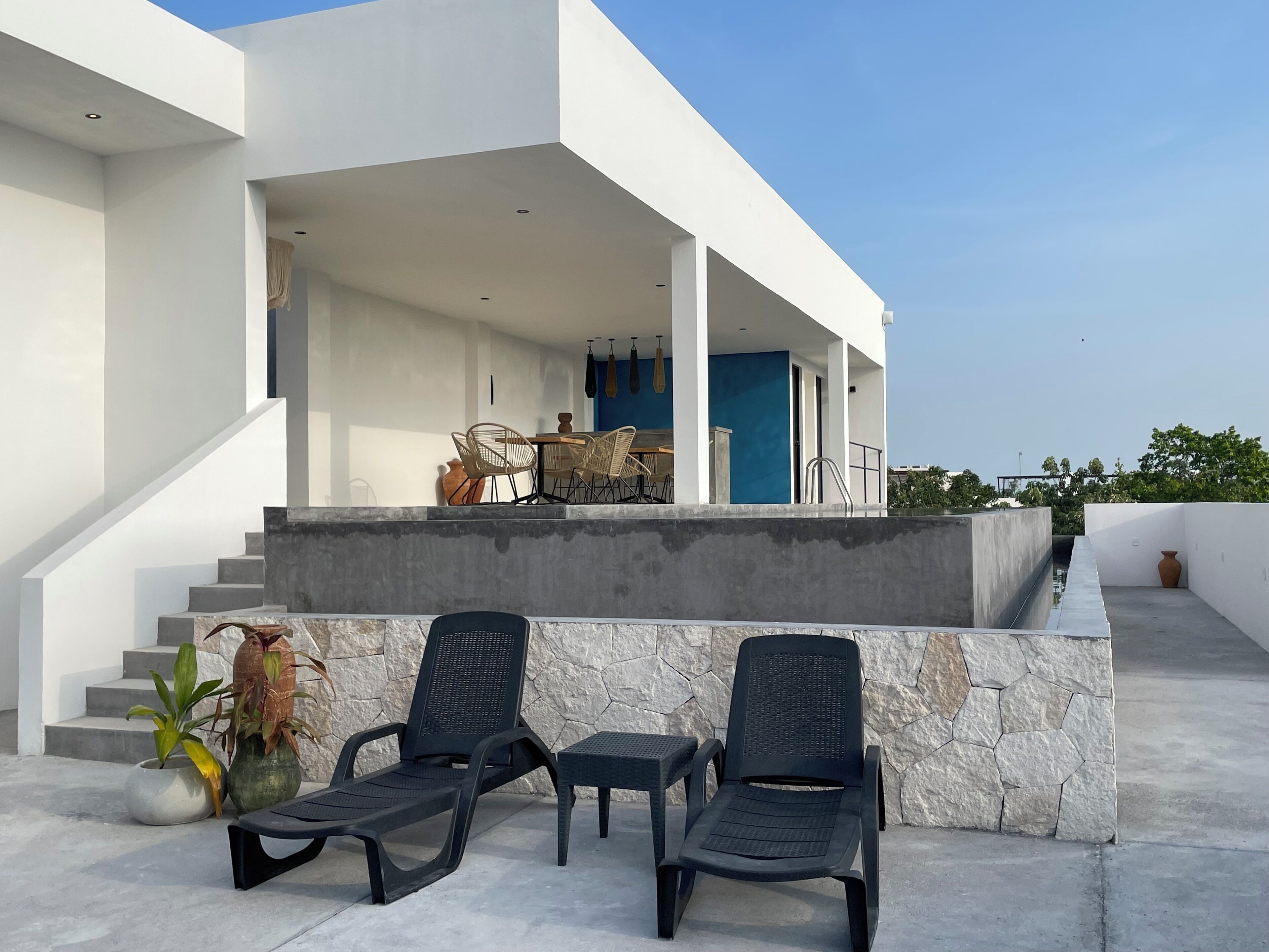 Un área de estar moderna al aire libre con dos sillones y una mesa, una escalera que lleva a un nivel superior y una vista del cielo.