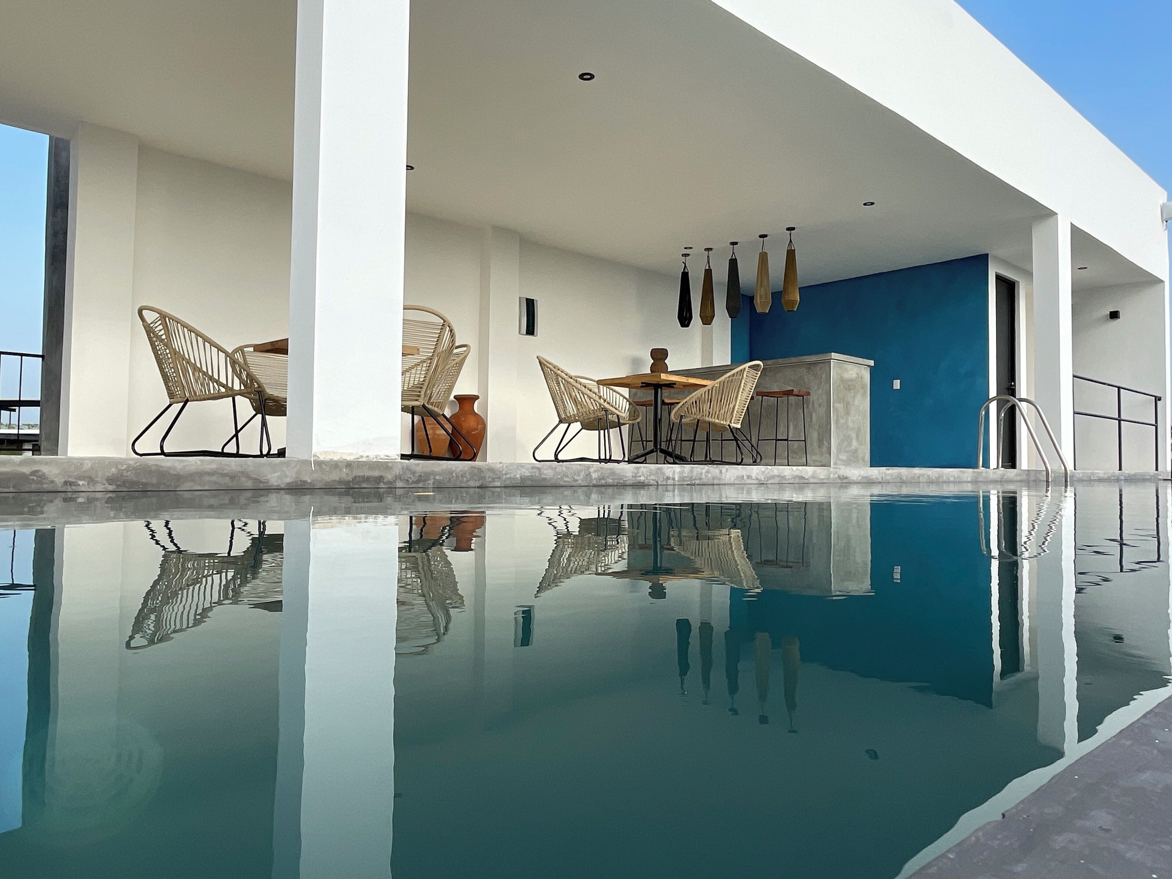Un área de piscina moderna al aire libre con agua cristalina, rodeada de paredes blancas y detalles azules. Se observan sillas de mimbre y una mesa con un florero.