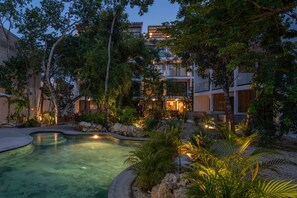 A pool area with a curved design, surrounded by tropical plants and trees, and adjacent to a multi-story building with large windows. - Erena Tulum (Tulum)