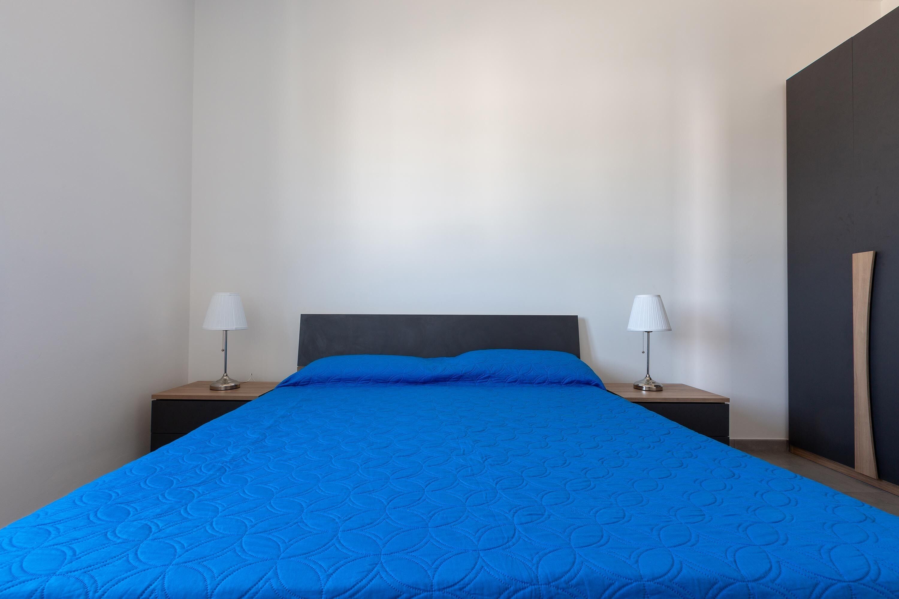 Un lit bien fait, avec une couverture bleue, deux lampes de chevet et une tête de lit sombre.