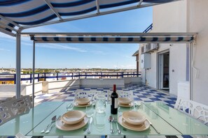 A glass table set for four with plates, cutlery, and a wine bottle, under a blue-striped awning, overlooking a coastal view. - 2505 Attico Primavera by Barbarhouse (Porto Cesareo)