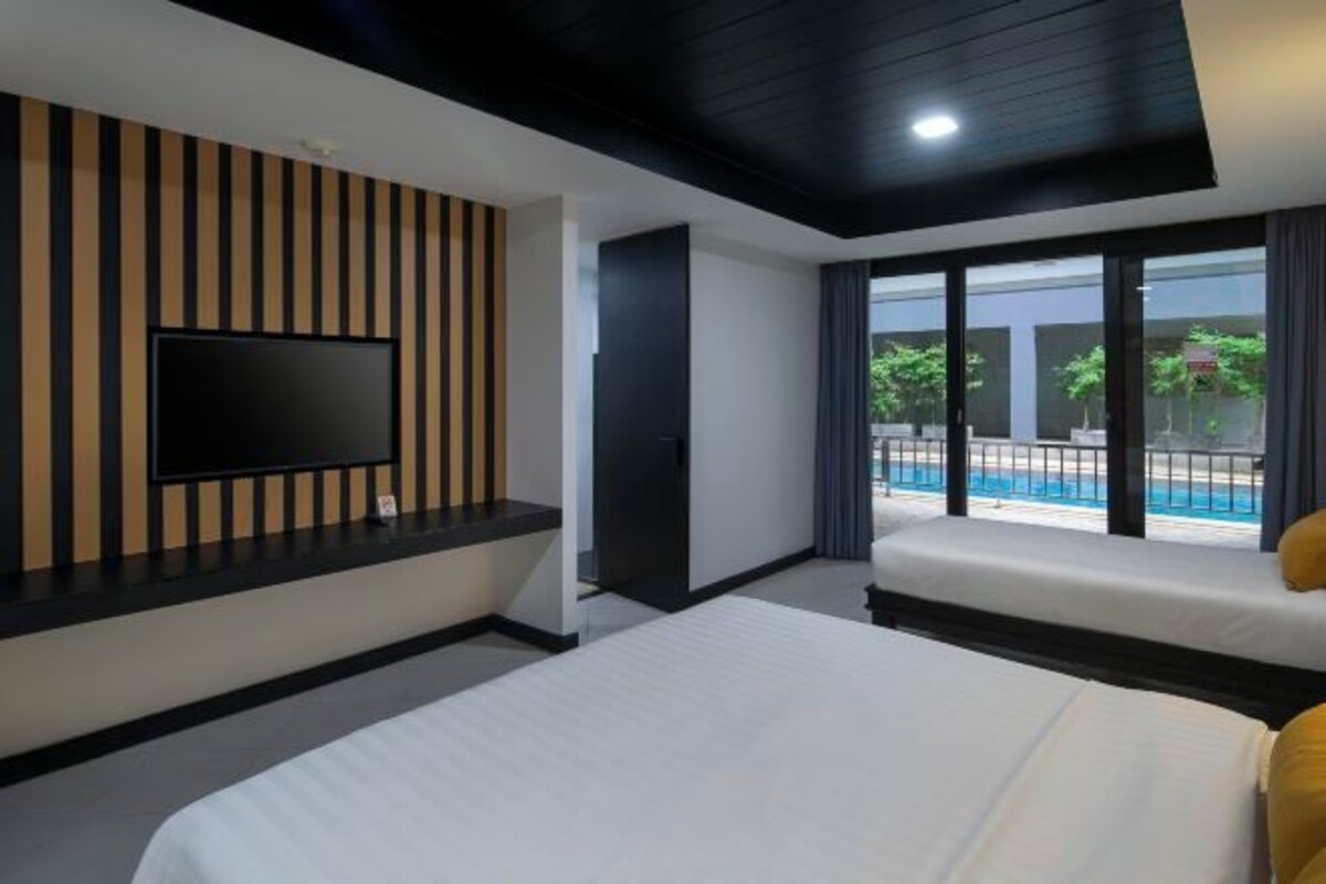Habitación de hotel moderna con una cama grande, un televisor de pantalla plana montado en una pared de paneles de madera, un balcón con vista a la piscina y un techo oscuro.