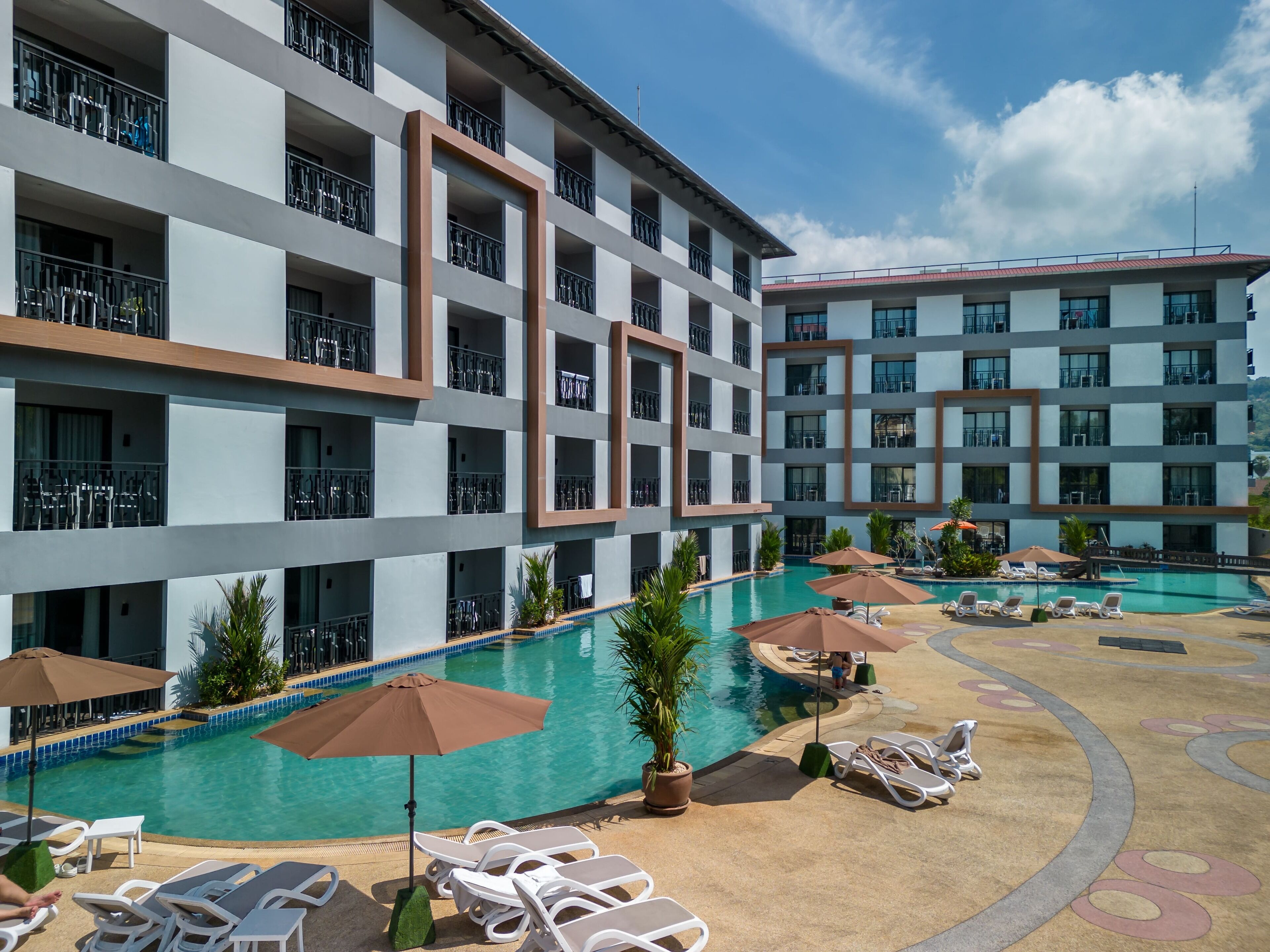 A hotel with multiple balconies, a swimming pool with sun loungers and umbrellas, and a clear blue sky.