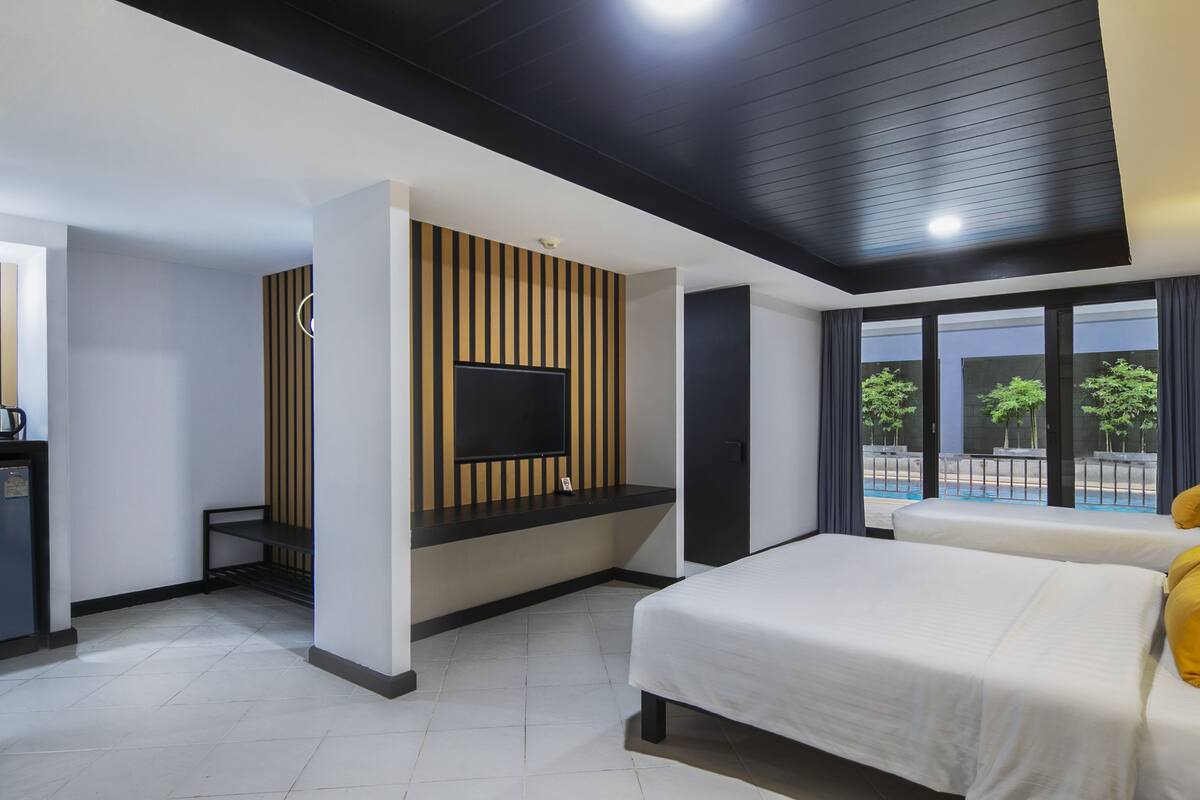 A modern hotel room with a large bed, a TV mounted on a wooden panel wall, a small desk with a kettle, and a view of a swimming pool through large windows.