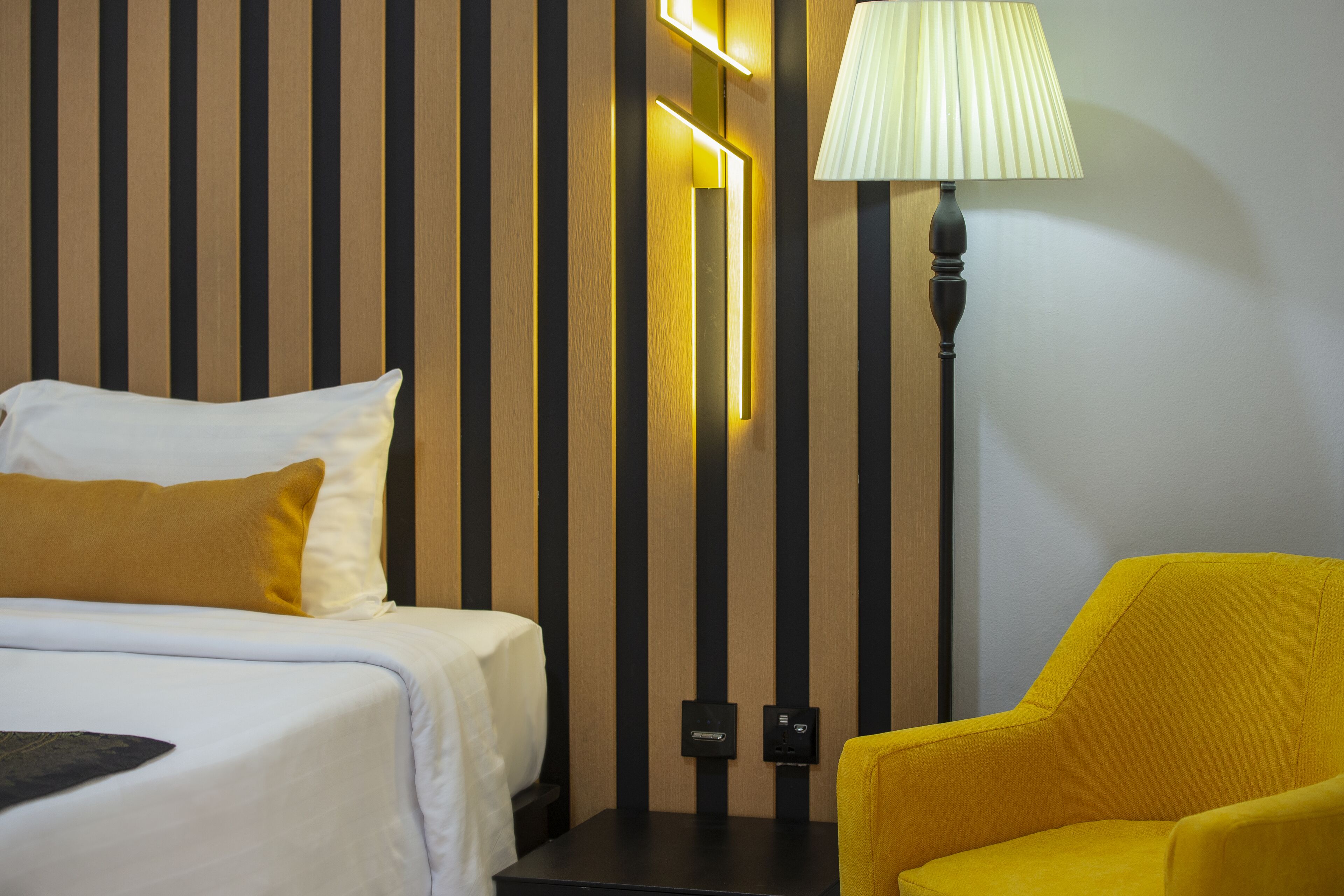 Una habitación de hotel con una cama, una mesita de noche, una lámpara y un sillón amarillo.
