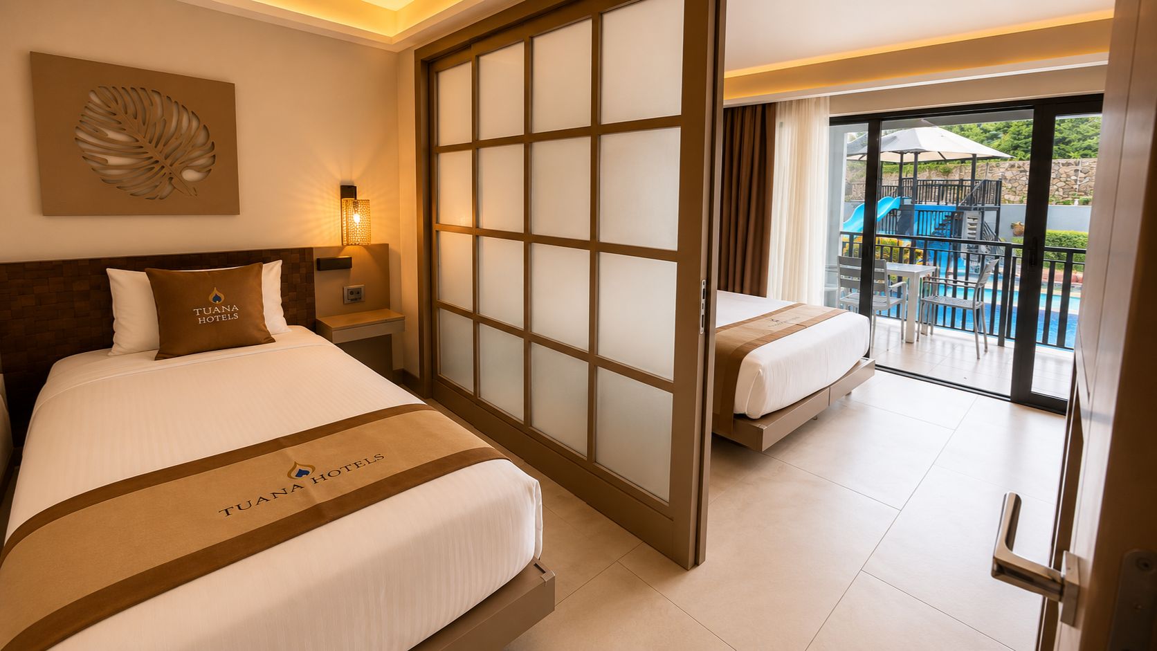 Habitación de hotel con cama, mesita de noche, lámpara de pared y puerta corredera de vidrio que da a un balcón con mobiliario exterior.