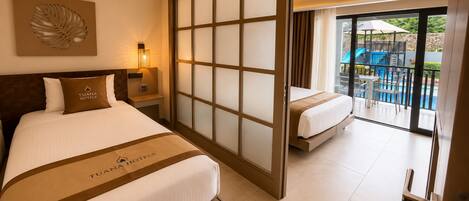Habitación de hotel con cama, mesita de noche, lámpara de pared y puerta corredera de vidrio que da a un balcón con mobiliario exterior.