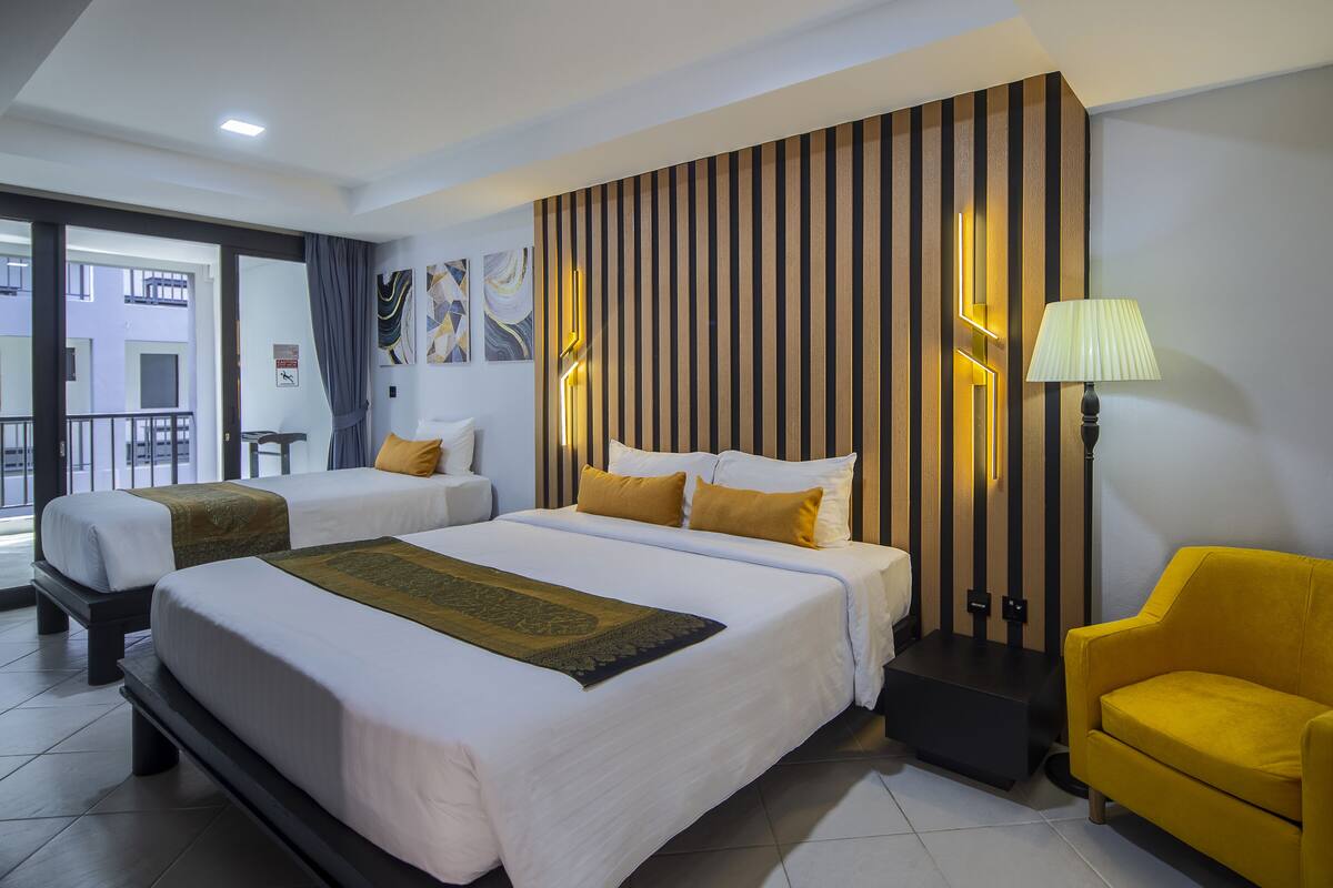 A hotel room with two beds, a yellow armchair, a standing lamp, and artwork on the wall.