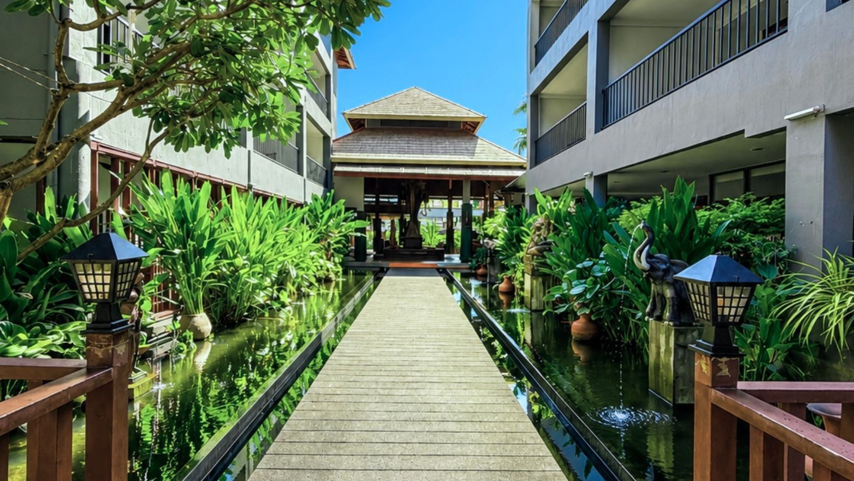 A corridor with a wooden walkway flanked by water features and greenery, leading to a building with a tiled roof.